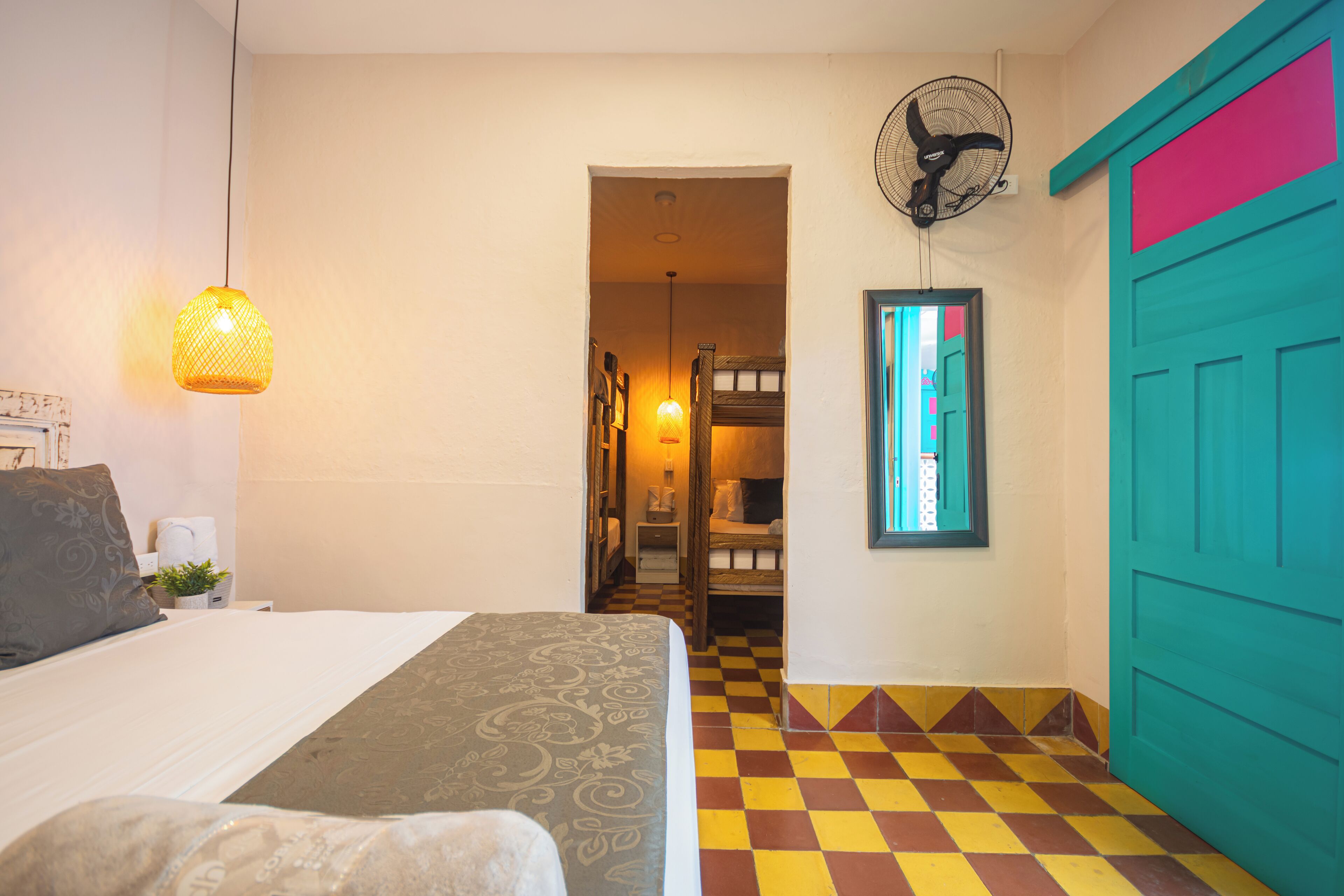 A hotel room with a bed, a hanging lamp, a fan, a checkered floor, and a teal door.