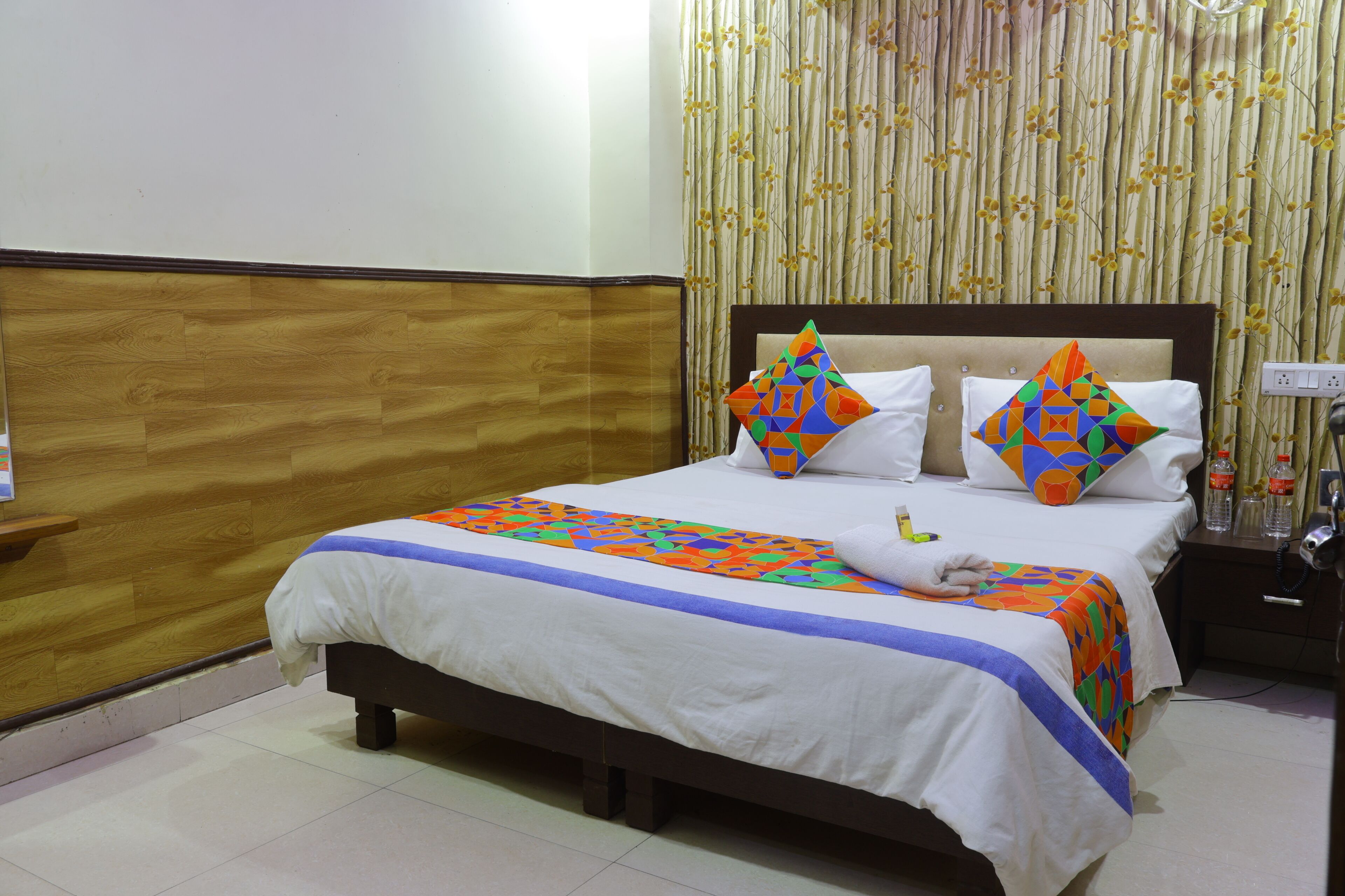 A hotel room with a wooden headboard, a bed with a colorful quilt and two decorative pillows, a bedside table with a bottle, and a curtain with a floral pattern.