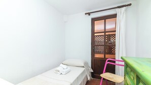 Ein kleines Zimmer mit einem Einzelbett, einer Holztür, einem grünen Schrank, einem rosa Stuhl und einem Fenster mit Blick ins Freie.