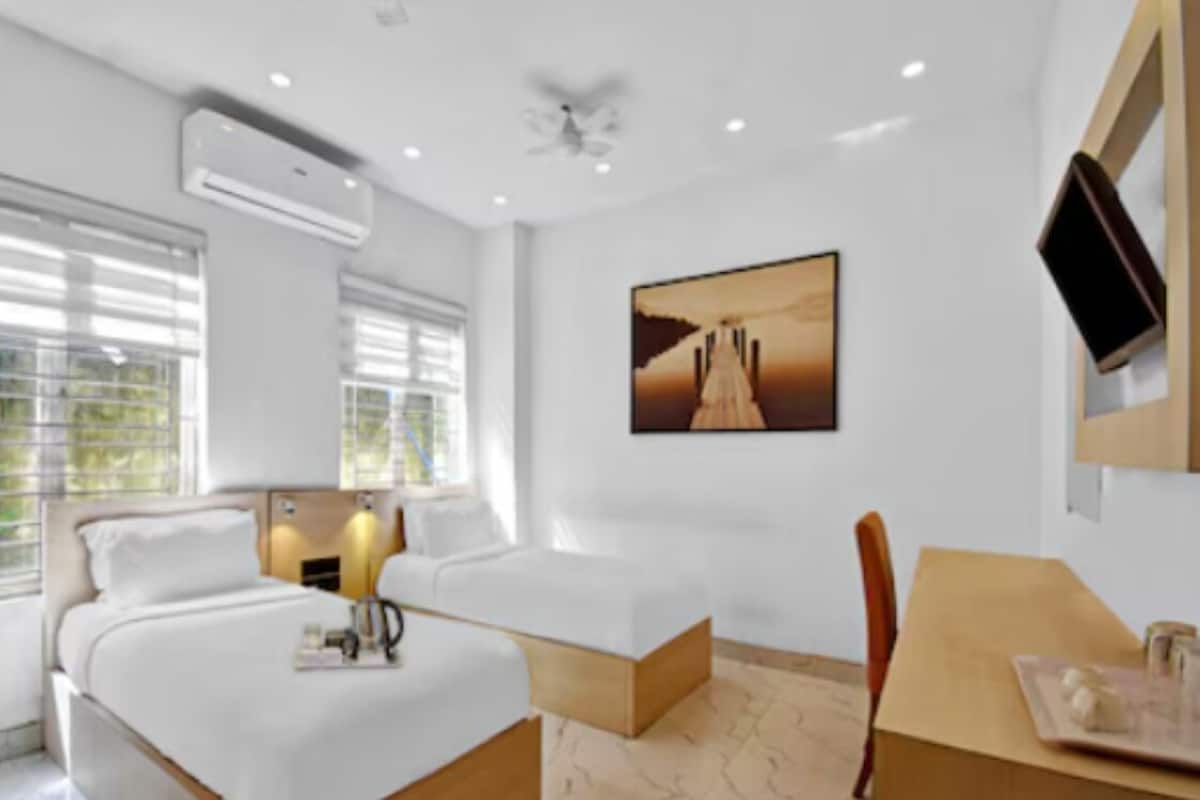A modern hotel room with a large bed, a desk, and a chair. There is a painting on the wall and an air conditioning unit on the ceiling.