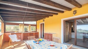 A covered patio with a table set for dining, wooden chairs, a window with curtains, and a sliding glass door leading to a balcony. - Trilocale H Ginepro Sgombro (Olbia)