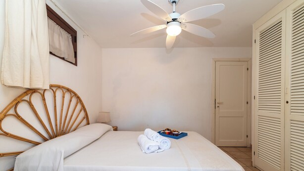 A neatly made bed with a white bedspread, a wooden headboard, and a ceiling fan. There is a tray with towels and a small bowl on the bed. - Trilocale H Ginepro Sgombro (Olbia)