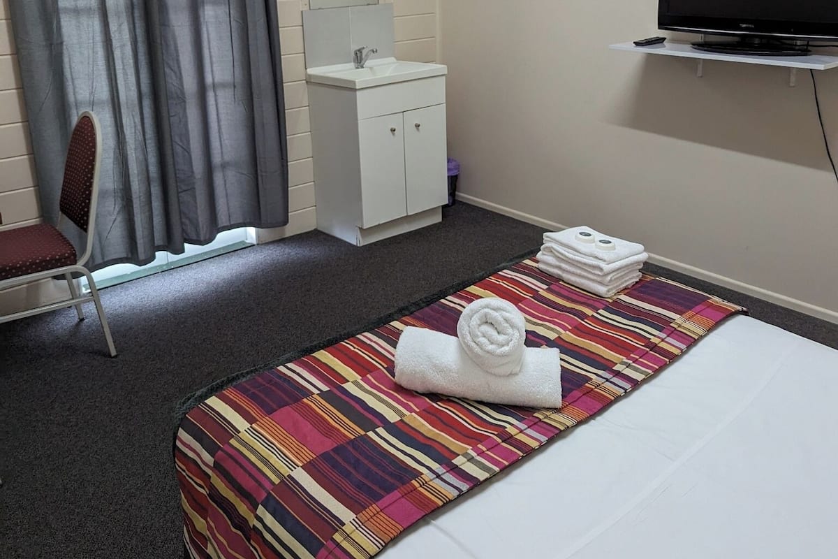 A hotel room with a bed, a television, a small white cabinet, a chair, and a window with curtains.