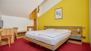 A room with a bed, desk, chair, and a small table with a lamp.