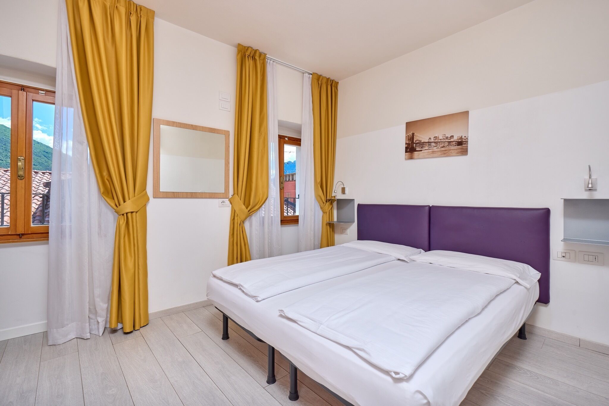 Une chambre à coucher avec un lit, des rideaux, un miroir, un tableau et une fenêtre donnant sur un beau paysage.