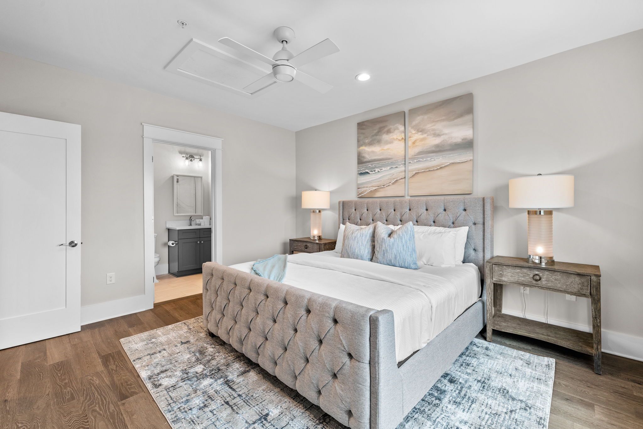 A bedroom with a tufted headboard bed, two bedside tables with lamps, a large painting on the wall, and a door leading to a bathroom.