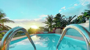 Une piscine avec des échelles en métal, entourée de palmiers et un coucher de soleil en arrière-plan.