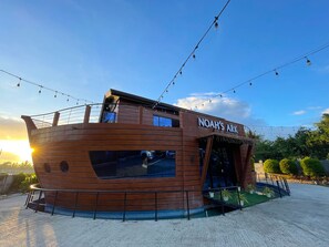 Un bâtiment en bois en forme de bateau, portant l’inscription « Arche de Noé » sur le côté, doté de grandes fenêtres et d’une entrée abritée.