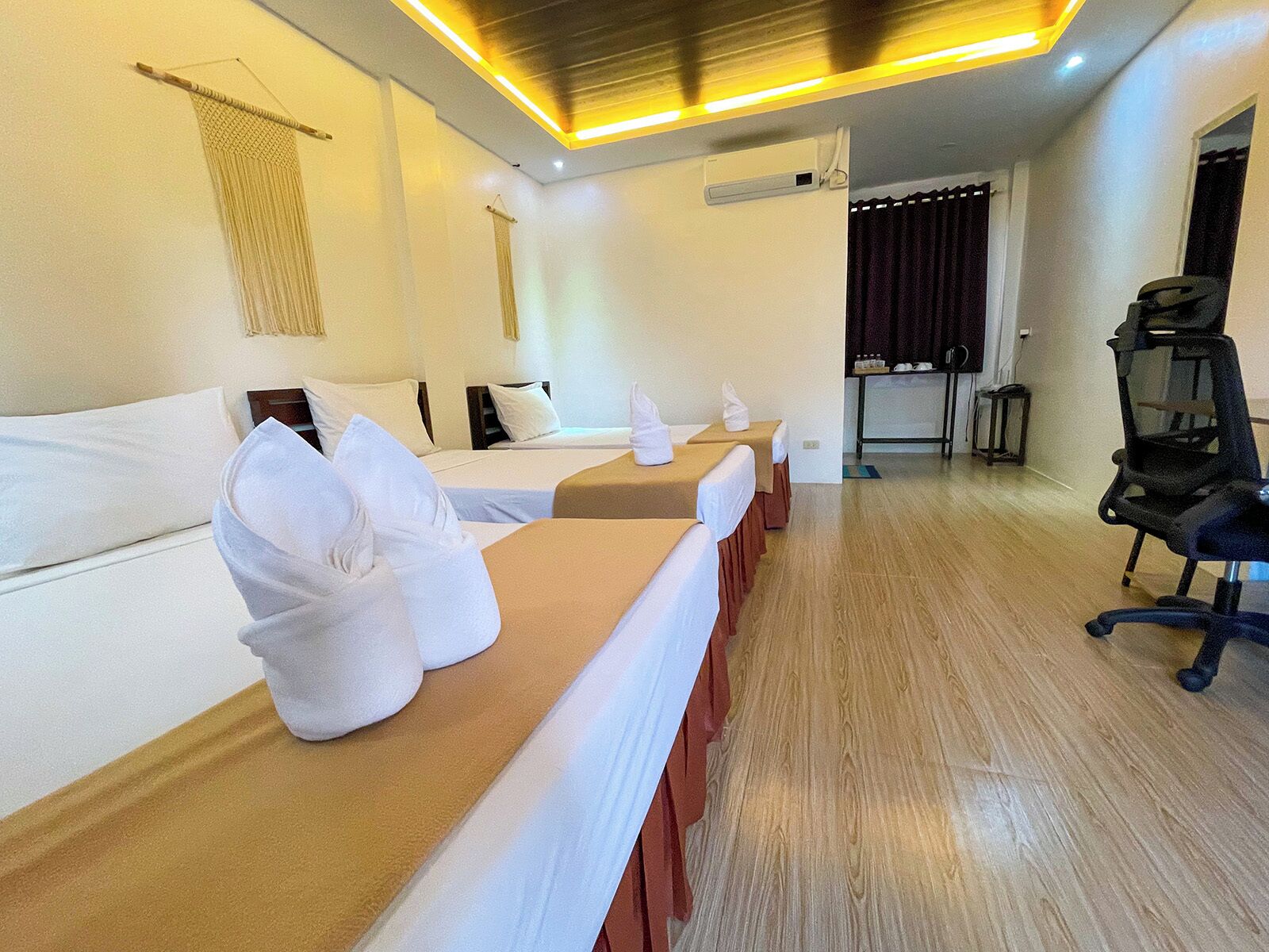 Quarto de hotel com três camas, uma escrivaninha com toalhas, uma cadeira e uma pequena mesa.
