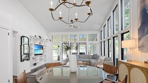 A spacious dining area with a round glass table surrounded by wooden chairs, a modern chandelier, and a large window offering a view of the outdoors. - 4 BR IOP Beach Rental with Private Pool & Hot Tub (Isle of Palms)