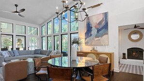 A spacious living room with a large round table, a fireplace, and a large window overlooking greenery. - 4 BR IOP Beach Rental with Private Pool & Hot Tub (Isle of Palms)