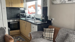 A compact kitchen with a stove, microwave, and sink, adjacent to a sofa with a plaid cushion. - 2 Bed Chalet Sleeps 4- Garden, Pets, Beach (Bridlington)