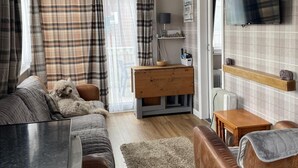 A cozy living room with a sofa, a wooden dresser, a TV mounted on the wall, and a window with curtains. - 2 Bed Chalet Sleeps 4- Garden, Pets, Beach (Bridlington)