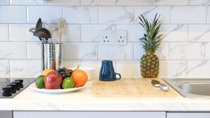 A kitchen with white marble tiles, a fruit bowl with various fruits, a blue mug, a pineapple, and a wooden cutting board. - Wonderful Holiday Studio in the Trendy Brick Lane (London)
