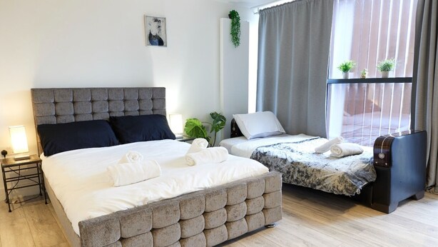 A double room with a large bed, a smaller bed, a bedside table, a lamp, and a window with curtains. - Wonderful Holiday Studio in the Trendy Brick Lane (London)