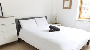 A neatly made bed with white linens, a bedside table with a lamp, and a view of the city through a window. - Apartment With Canal View in Broadway Market (London)