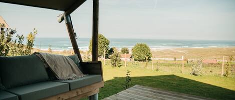 A wooden deck with a green sofa and a small table, overlooking a grassy area with trees and a view of the ocean.