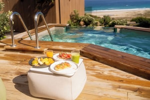 Una terraza de madera con piscina, una mesa con platos de comida y un vaso de jugo, y vista a la playa.