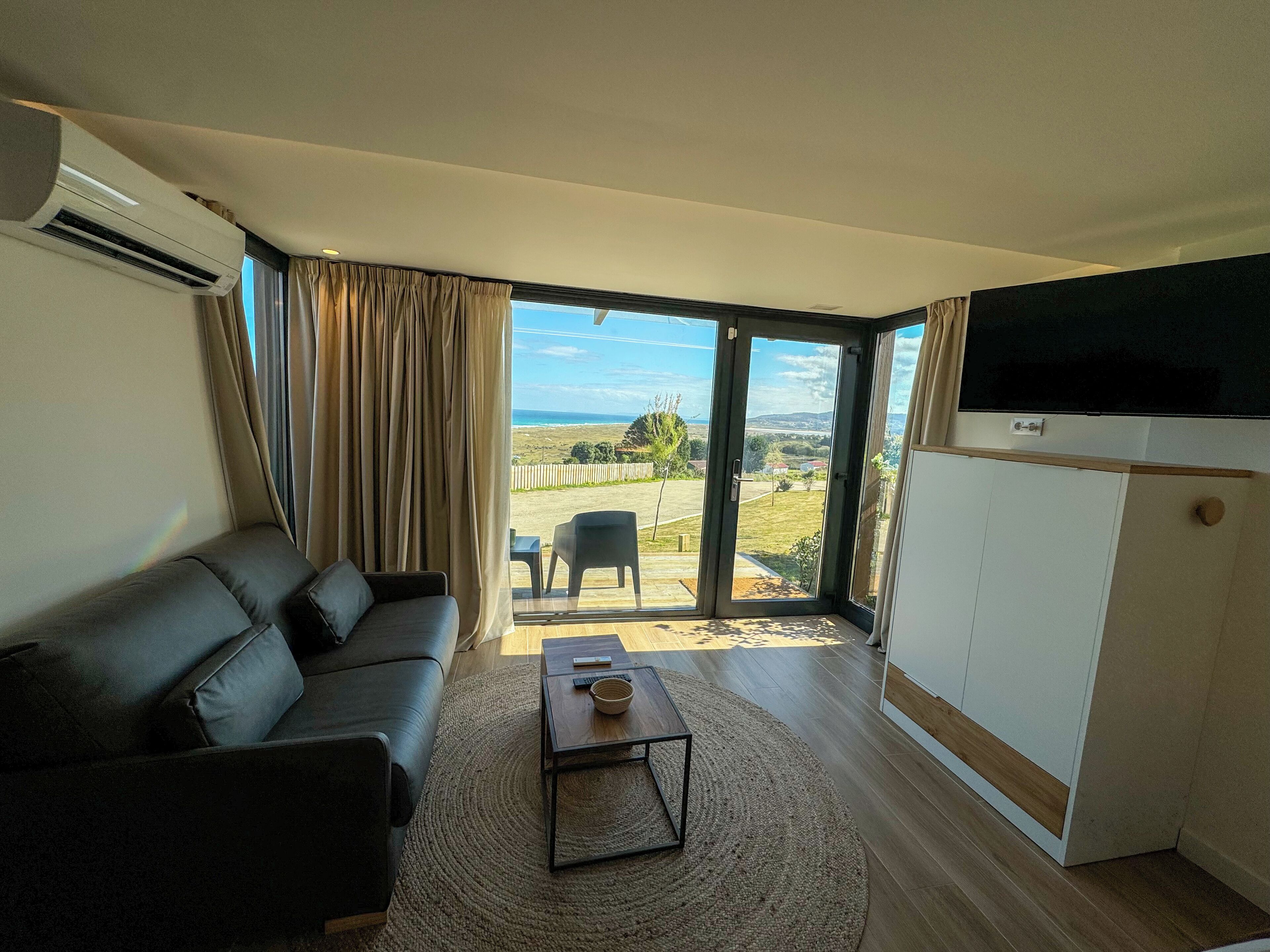 A modern living room with a black leather sofa, a wooden coffee table, and a large sliding glass door leading to an outdoor area with a view of the ocean.