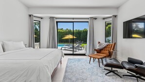 Un dormitorio moderno con una cama grande, un espacio de descanso con sillas y vistas a una piscina y vegetación a través de un ventanal corredero.