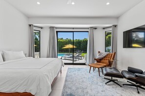 A modern bedroom with a large bed, a seating area with chairs, and a view of a pool and greenery through a sliding glass door.
