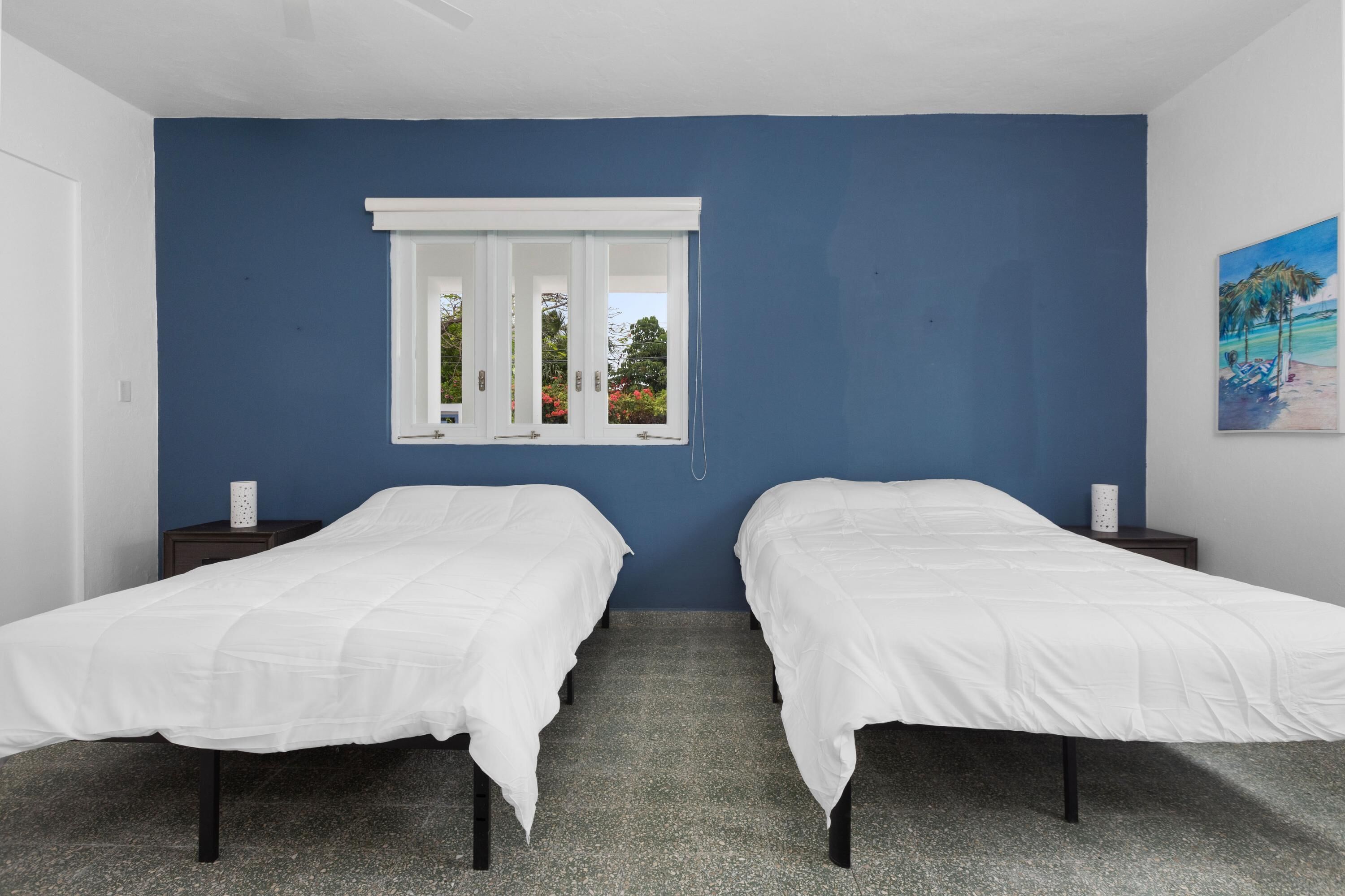 A room with two single beds, a window with white shutters, a small nightstand with a lamp, and a painting on the wall.