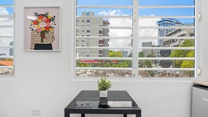 Ein Zimmer mit großem Fenster, einem gerahmten Kunstwerk an der Wand und einem kleinen Tisch mit einer Topfpflanze.