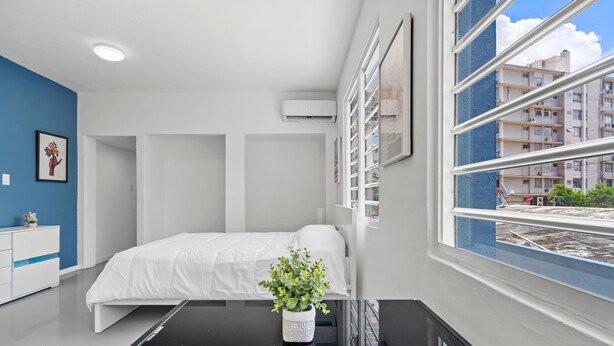 Ein modernes Schlafzimmer mit einem Bett, einer Zimmerpflanze, einer weißen Kommode und einem Fenster mit Blick auf Gebäude.