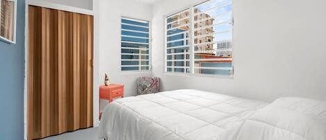 A bedroom with a large bed, a wooden door, a small table, and a window with blinds.