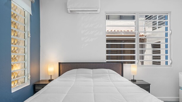 A bedroom with a large bed, two bedside lamps, a window with blinds, and an air conditioning unit on the wall. - Otium Rentals Condado Space Near Attractions (San Juan)