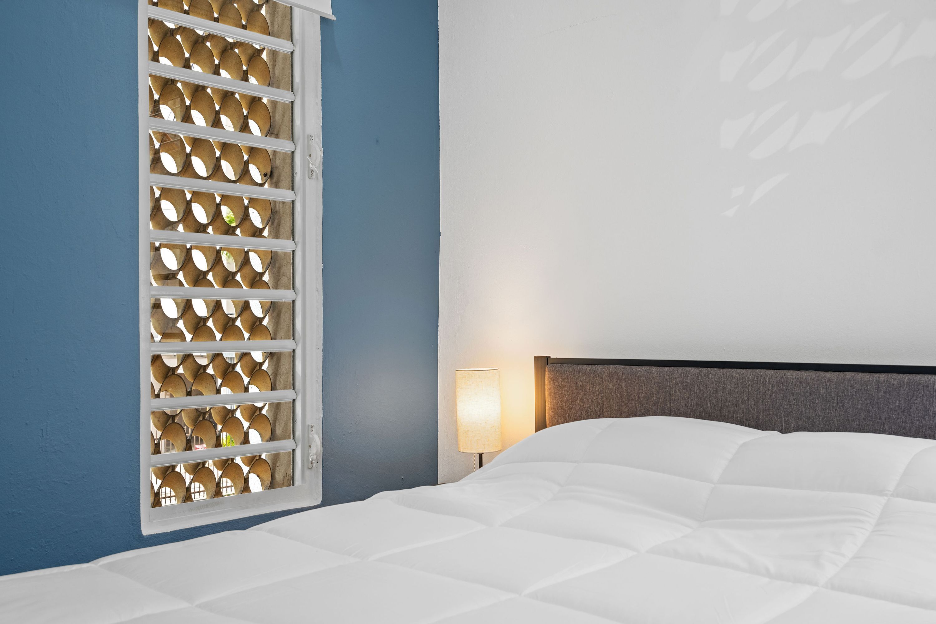 A hotel room with a bed, a window with a wooden lattice, and a wall lamp.