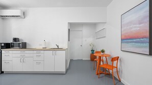 A modern kitchen with white cabinetry, a microwave, and a coffee maker. There is a dining area with orange chairs and a small table. A large painting of a sunset is on the wall. - Otium Rentals Condado Space Near Attractions (San Juan)