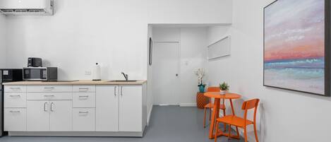A modern kitchen with white cabinetry, a microwave, and a coffee maker. There is a dining area with orange chairs and a small table. A large painting of a sunset is on the wall.