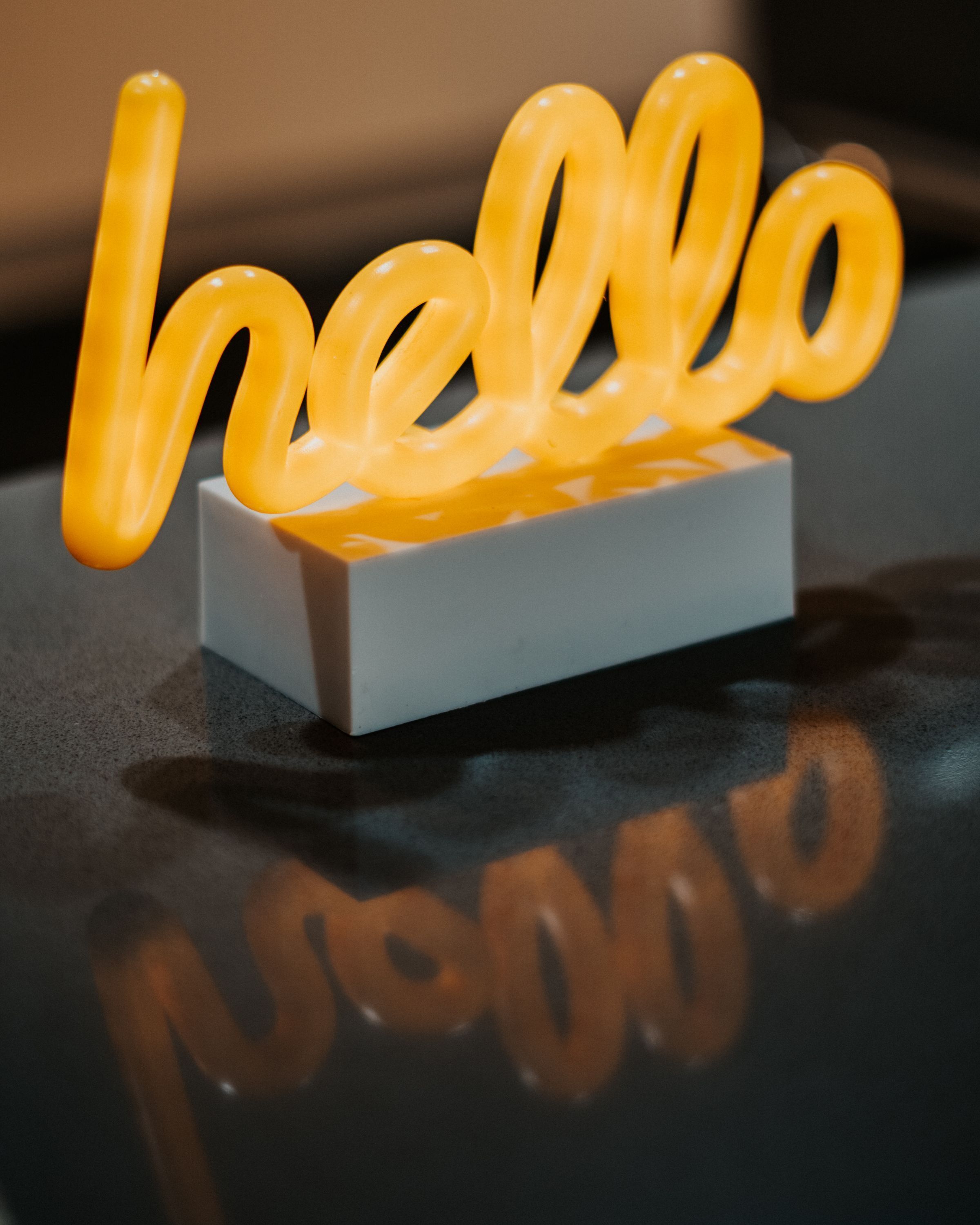 Un panneau lumineux au néon, affichant le mot « hello » dans une police stylisée, fixé sur un socle rectangulaire blanc et placé sur une surface réfléchissante.