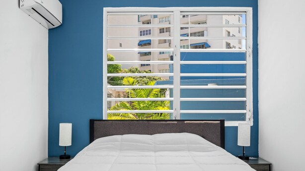 Une chambre d’hôtel avec un lit, une fenêtre avec des stores, une table de chevet et un climatiseur fixé au mur.