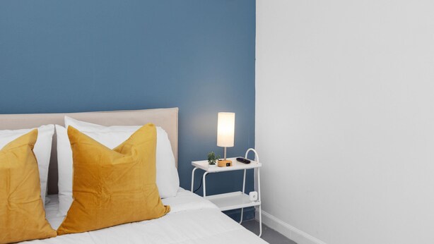 A neatly made bed with white and mustard yellow pillows, a bedside table with a lamp, and a small plant. - Otium Rentals Homely Haven in Condado (San Juan)