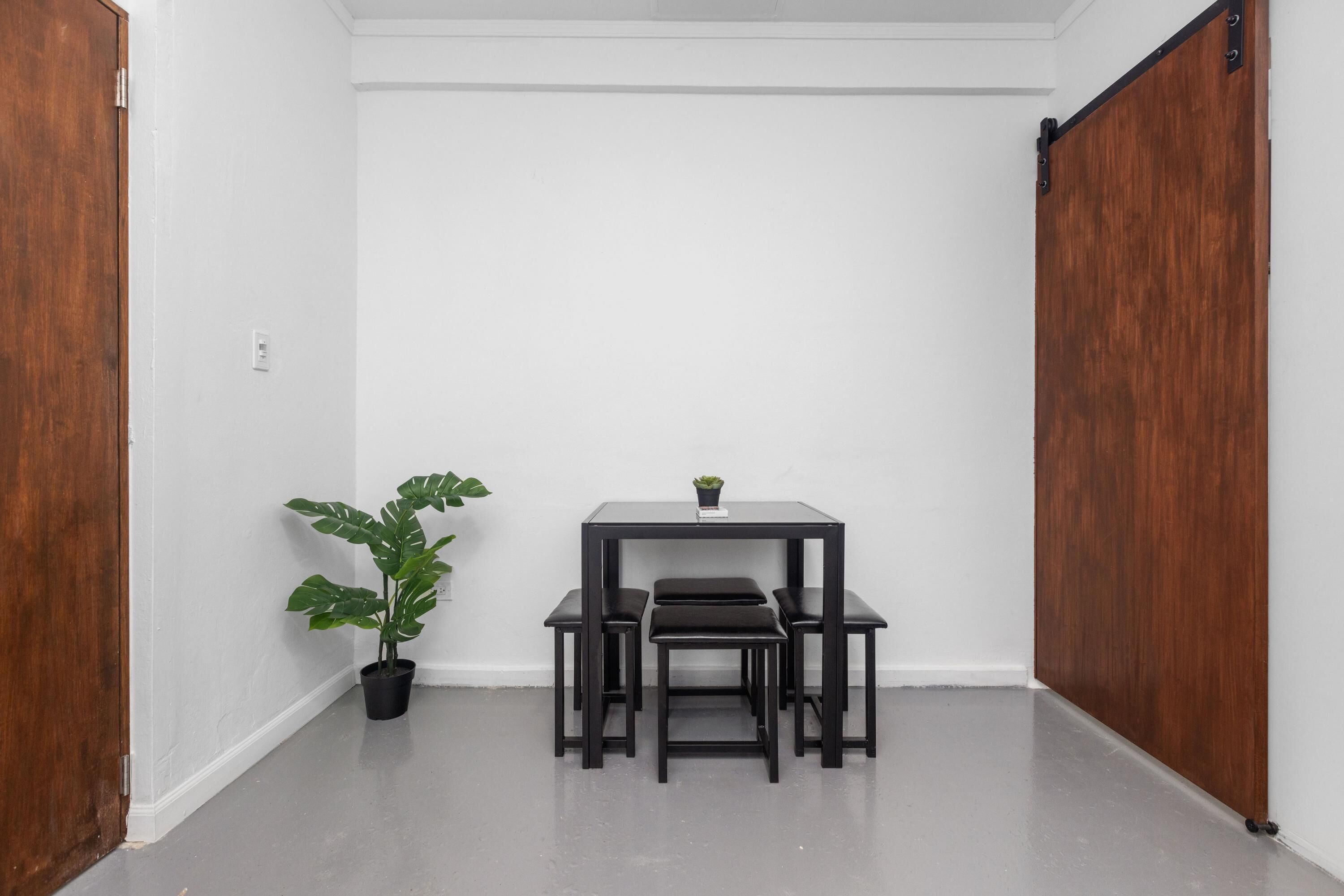 A corner room with a wooden door, white walls, a black table with a bench, and a potted plant.