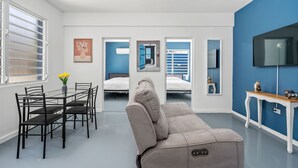 A modern room with a sofa, dining table, chairs, and a TV. There are two beds in the background. - Otium Rentals Ideal Space for Familes Friends (San Juan)