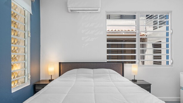 A bedroom with a bed, two bedside lamps, an air conditioner, a window with blinds, and a wardrobe with a mirror. - Otium Rentals Ideal Space for Familes Friends (San Juan)