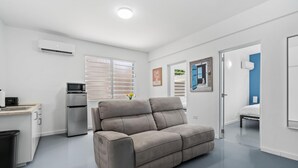 A modern, well-lit room with a grey sofa, a kitchenette with a fridge and microwave, a window with blinds, and a door leading to another room. - Otium Rentals Ideal Space for Familes Friends (San Juan)