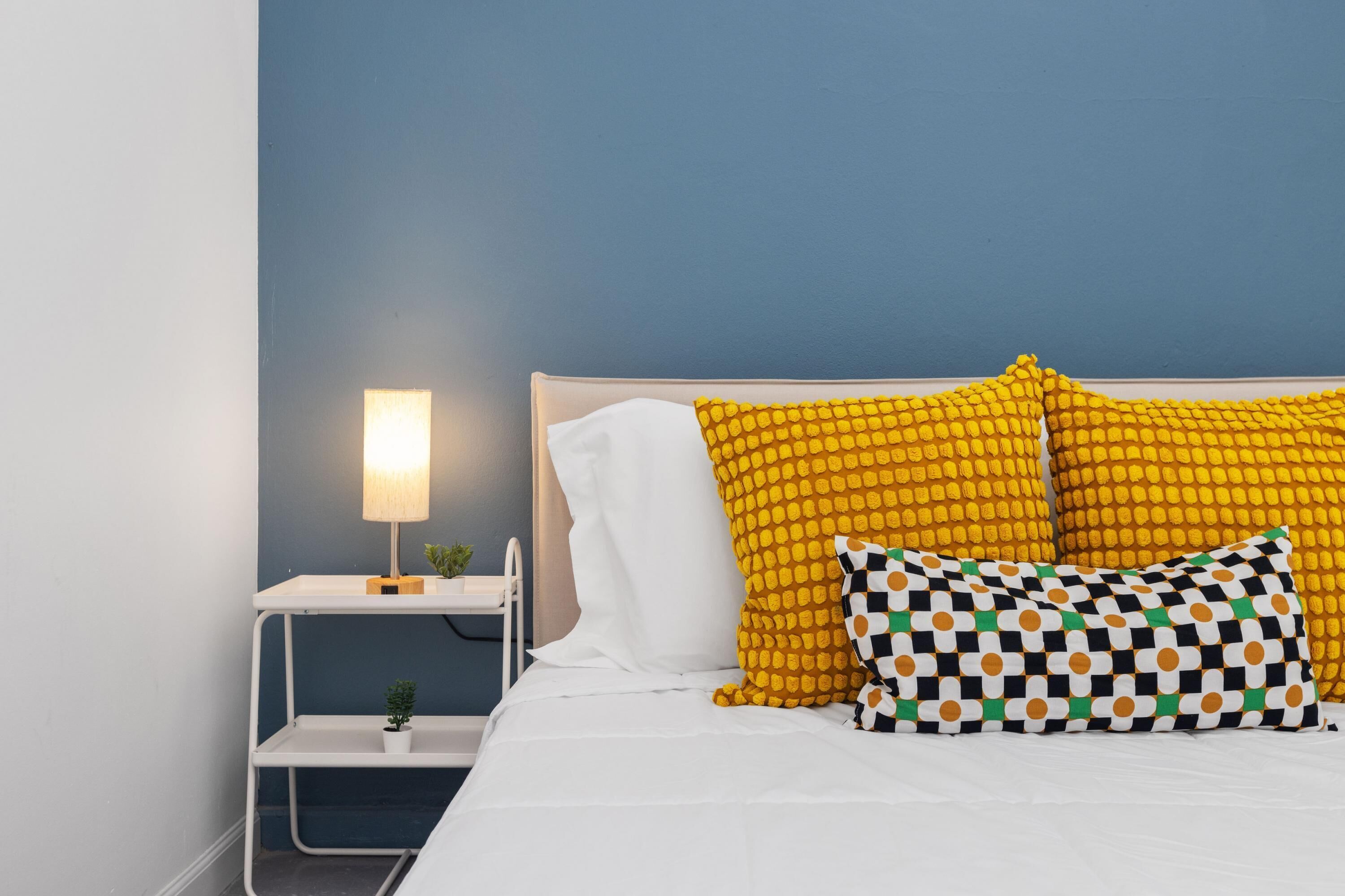 A bed with white bedding, a blue wall, and a nightstand with a lamp and a small plant.