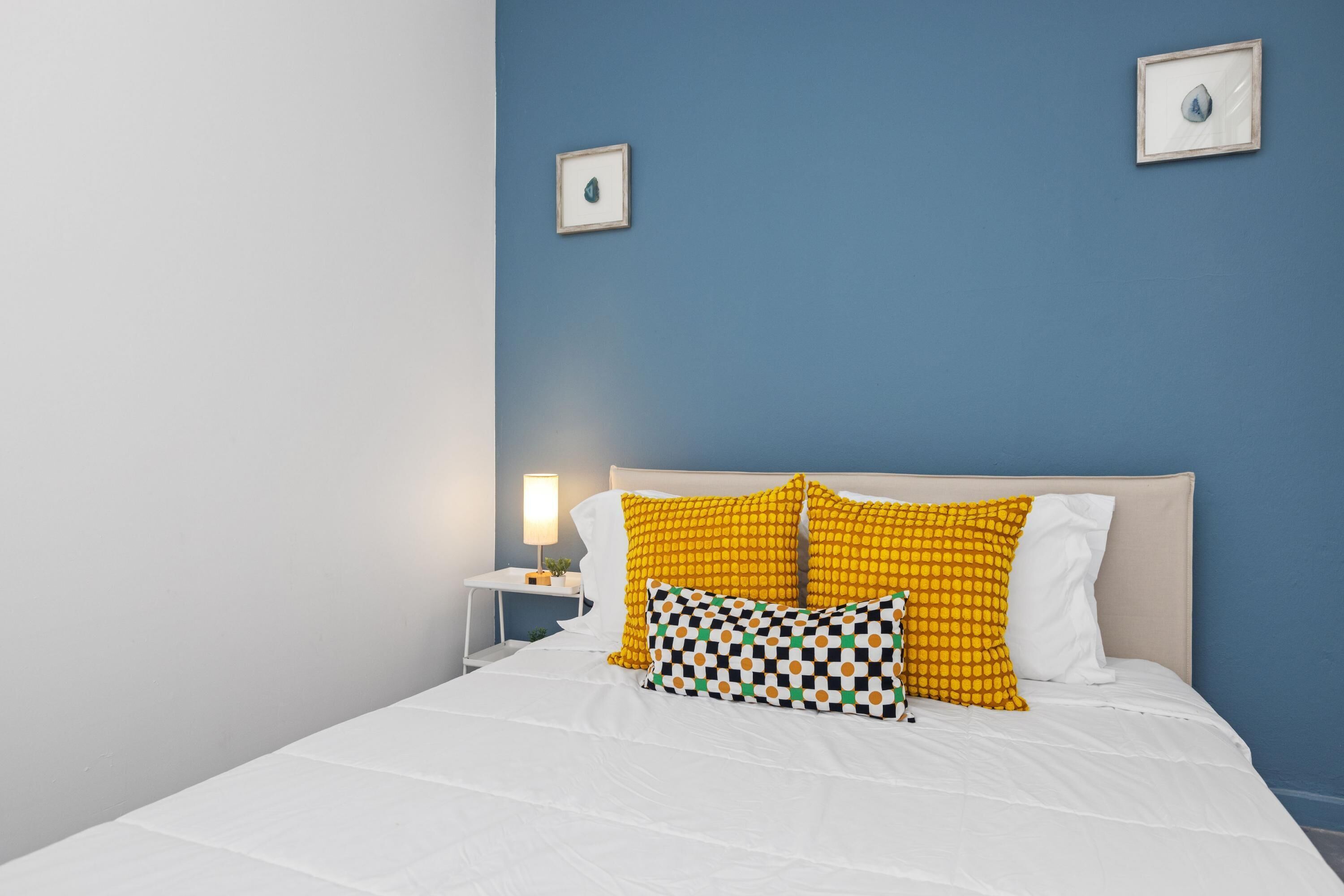 Una cama bien hecha con ropa de cama blanca y dos almohadas, una amarilla y otra estampada con cuadros, frente a una pared azul.