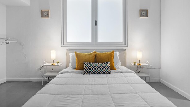 Quarto com cama grande, duas mesas de cabeceira com luminárias, janela com persianas e um pequeno quadro na parede.
