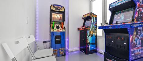 A room with two arcade machines, a white folding chair, and a window with natural light.