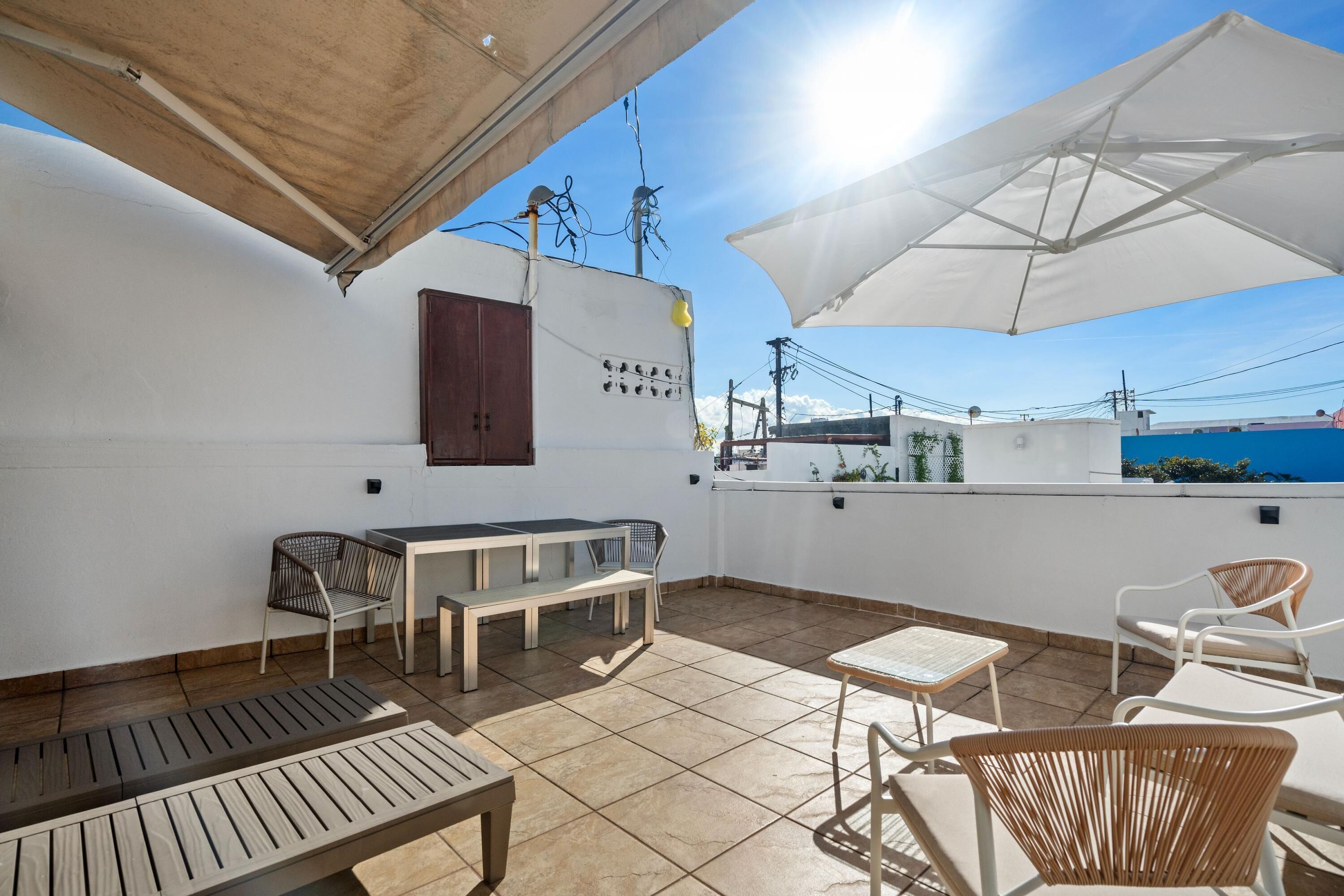 Une terrasse extérieure avec une table et des chaises, un banc en bois et un mur blanc orné d’un objet décoratif.