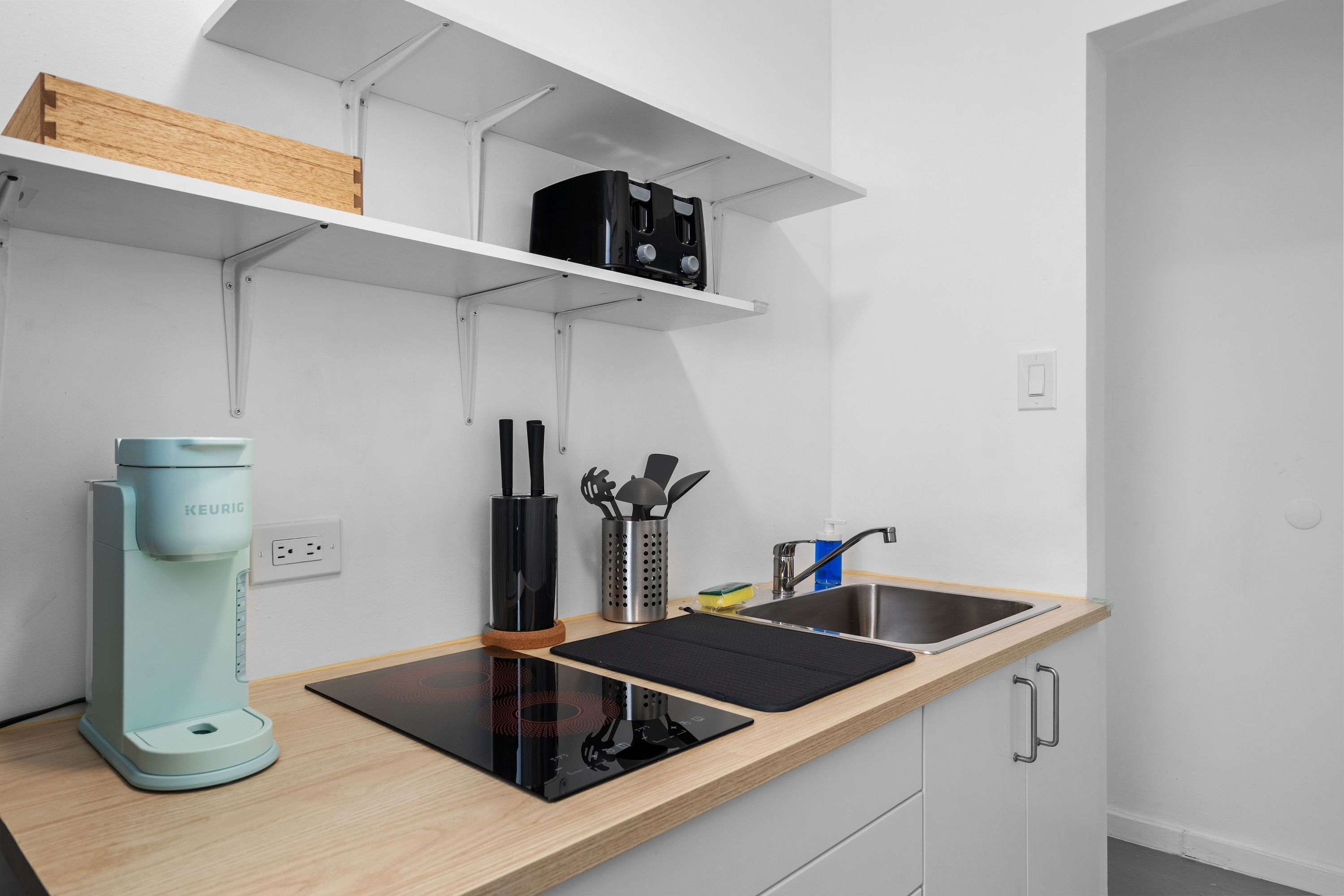 A modern kitchen with a built-in coffee maker, a black induction cooktop, a sink, and a wooden shelf above.