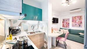 A compact kitchen with teal cabinets, a coffee machine, and a dining area with a green sofa and pink chair. - Urban Bloom Oasis - Mini Zen Maison (Kalamata)
