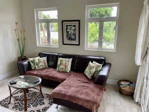 A living room with a dark leather sofa, a glass coffee table, and a potted plant. There are two windows with curtains, a framed picture on the wall, and a basket of books on the floor. - Remarkable 3-bed Villa in Christ Church (Providence)