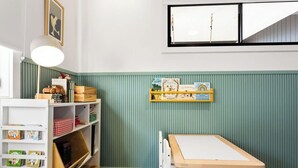Une chambre d’enfant avec une table blanche, des étagères remplies de livres et de jouets, et une fenêtre avec des stores.