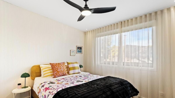 Une chambre d’hôtel avec un lit, un ventilateur de plafond, une fenêtre avec des rideaux et une table de chevet avec une lampe.